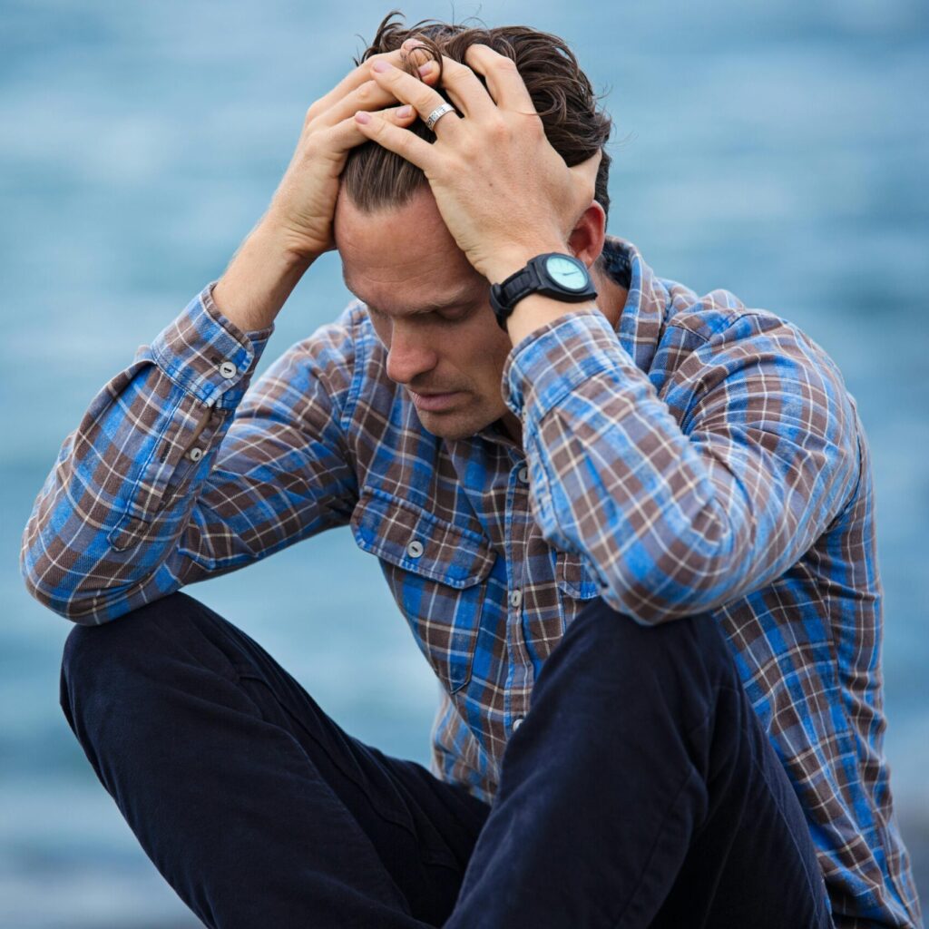 A man in a plaid shirt sits by the water looking distressed, symbolizing stress.